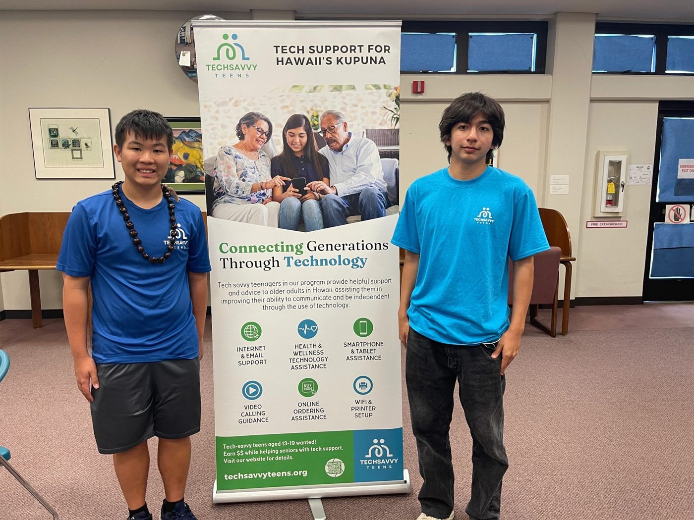 Two teen volunteers beside a Tech Savvy Teens banner reading Connecting Generations Through Technology